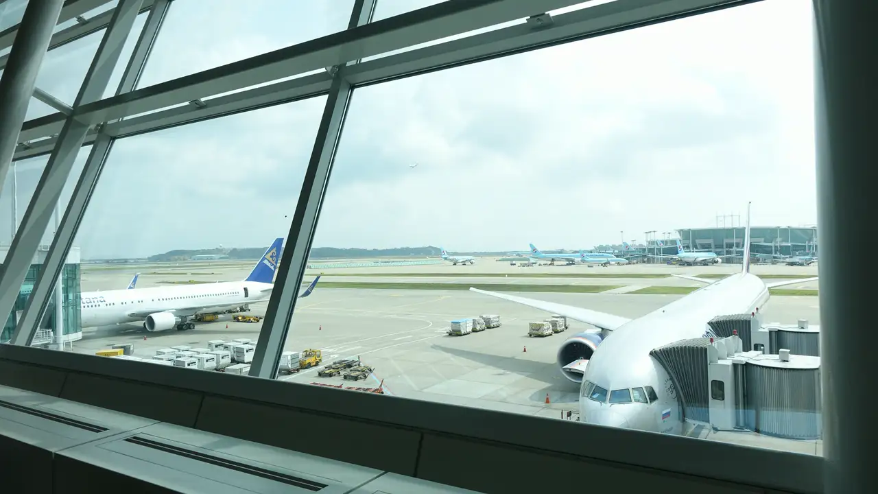 An airliner docked at a jet bridge and the airfield beyond, seen through the windows of a boarding gate area at Incheon International Airport