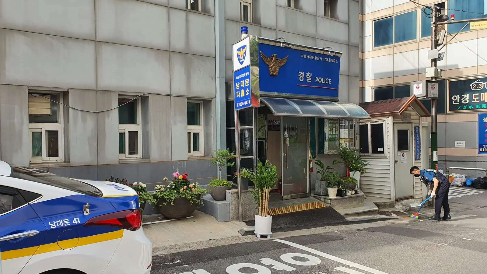 The front of Namdaemun Police Box in Seoul, with a blue Police sign over the door, potted plants by the entrance, a patrol car in the foreground and an officer sweeping the pavement beside the building