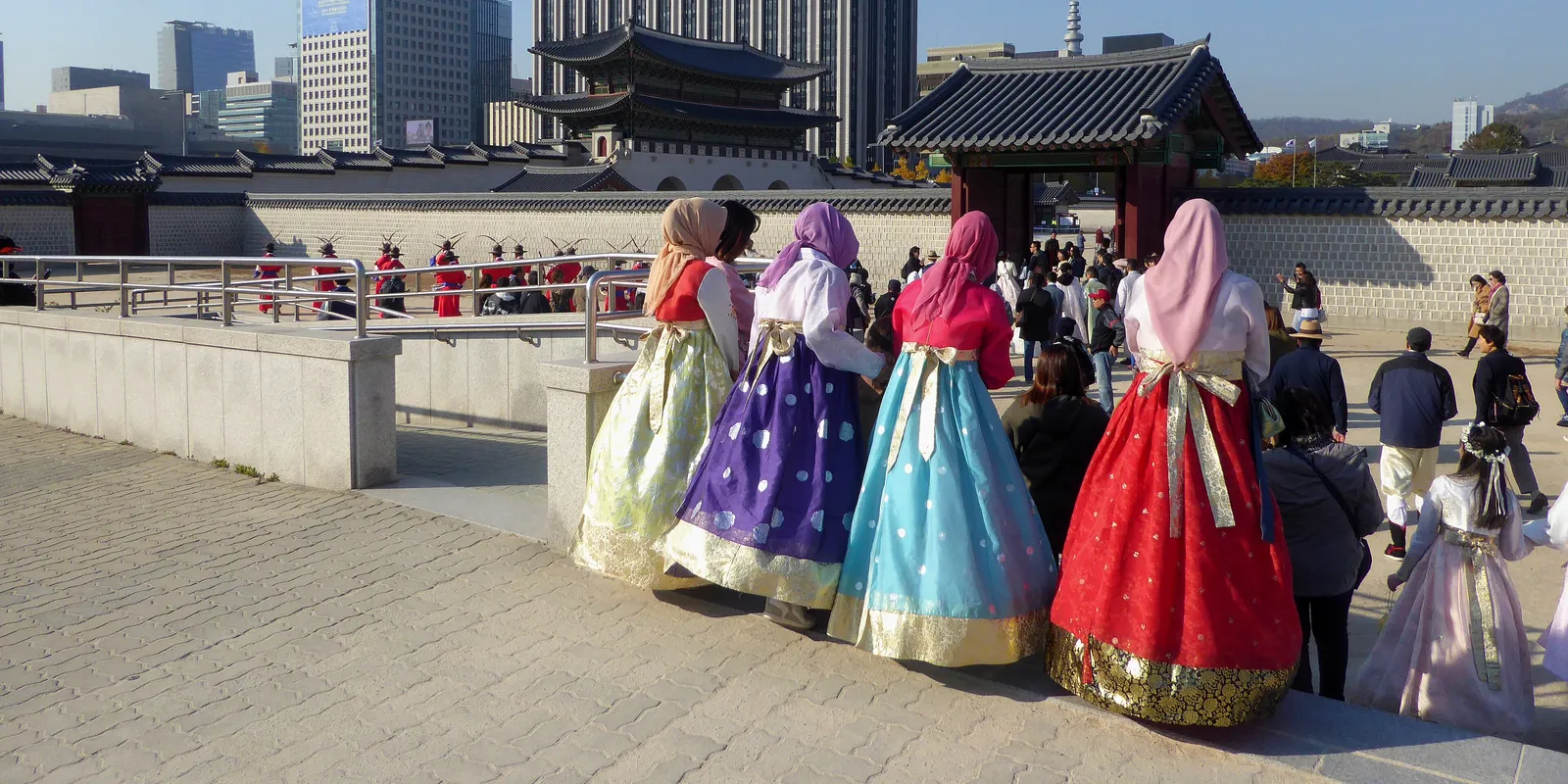 A group of travelers in hanbok seen from behind, standing side by side inside Gyeongbokgung Palace and watching guards in red uniforms