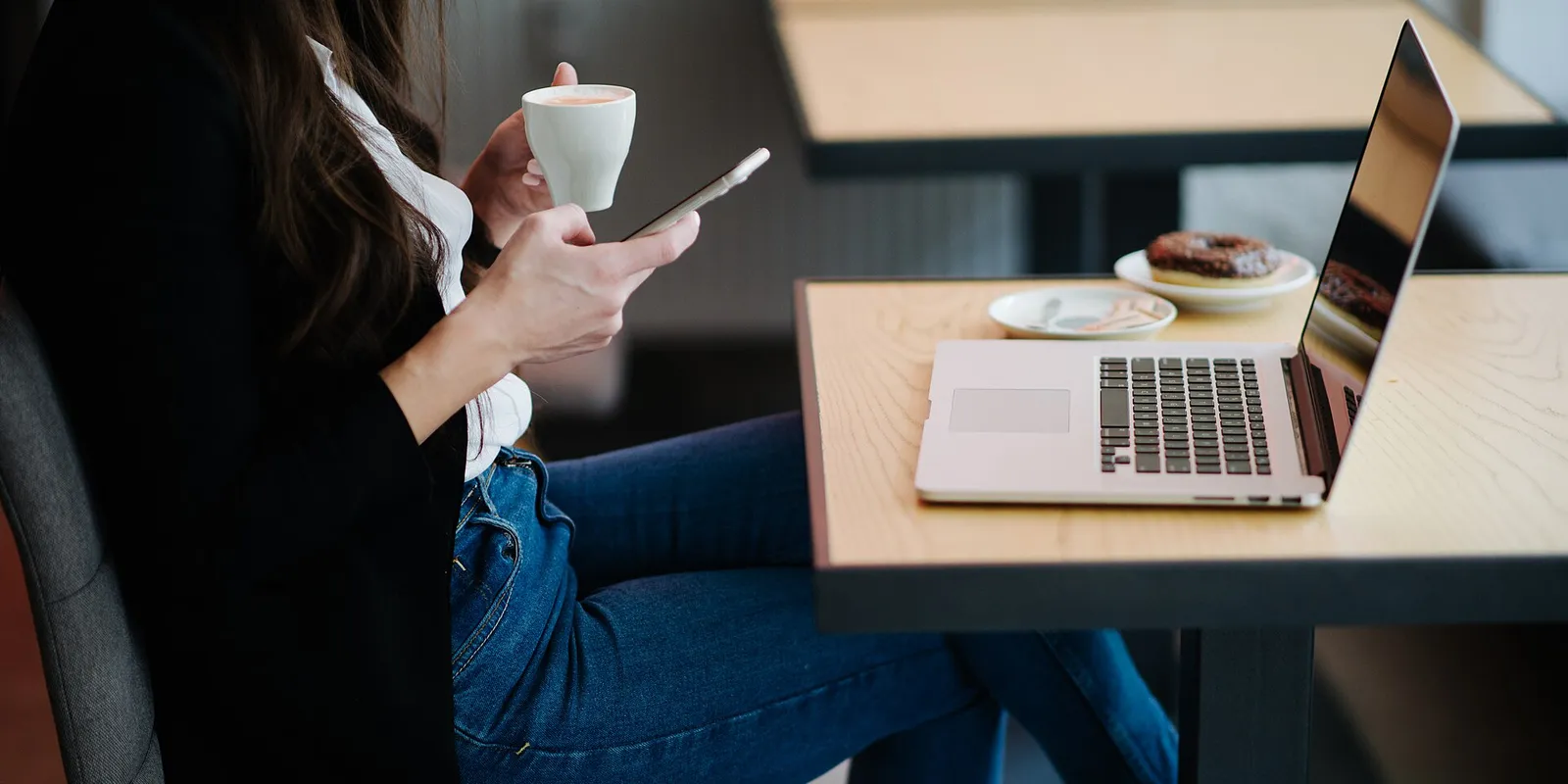 A person at a café table holding a coffee cup in one hand and a smartphone in the other, with an open laptop on the table