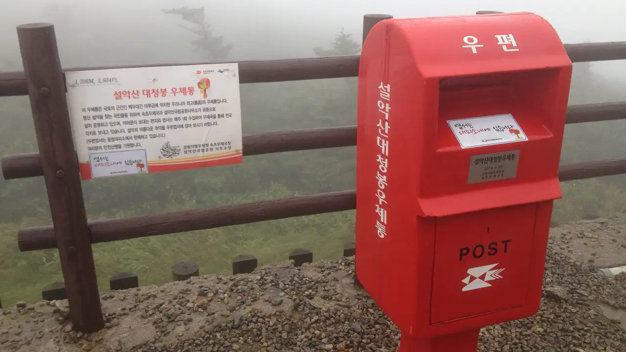 A red postbox on the fog-covered summit of Daecheongbong in Seoraksan, with an information sign on the railing beside it