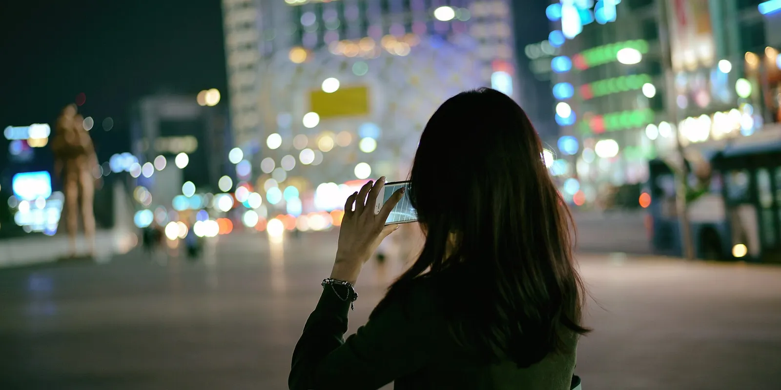 A person seen from behind, photographing city lights with a smartphone on a Seoul street at night