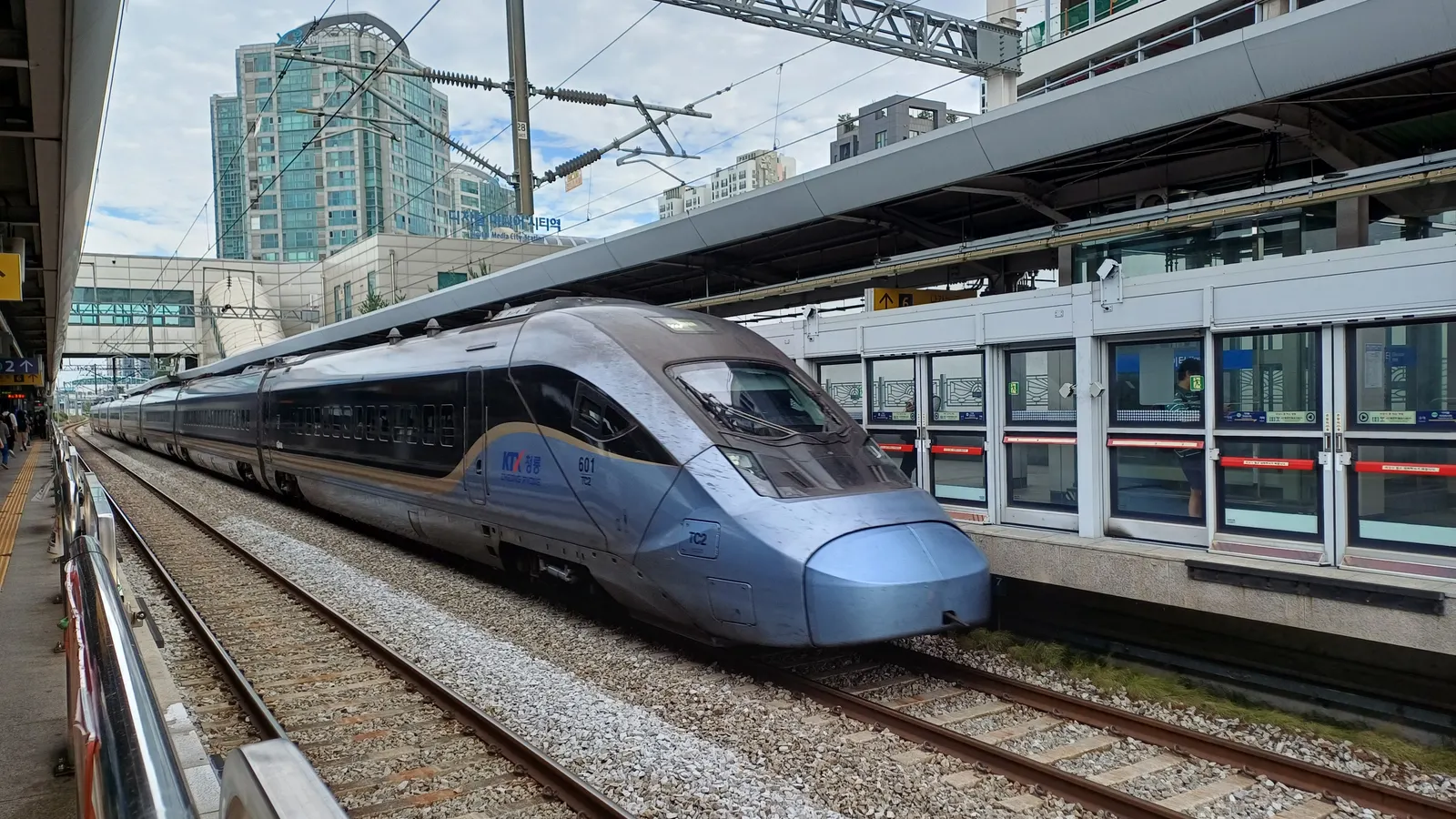 A blue-grey KTX-Cheongryong high-speed train running along the track beside the platform at Digital Media City Station in Seoul, with platform screen doors on the right and apartment towers behind