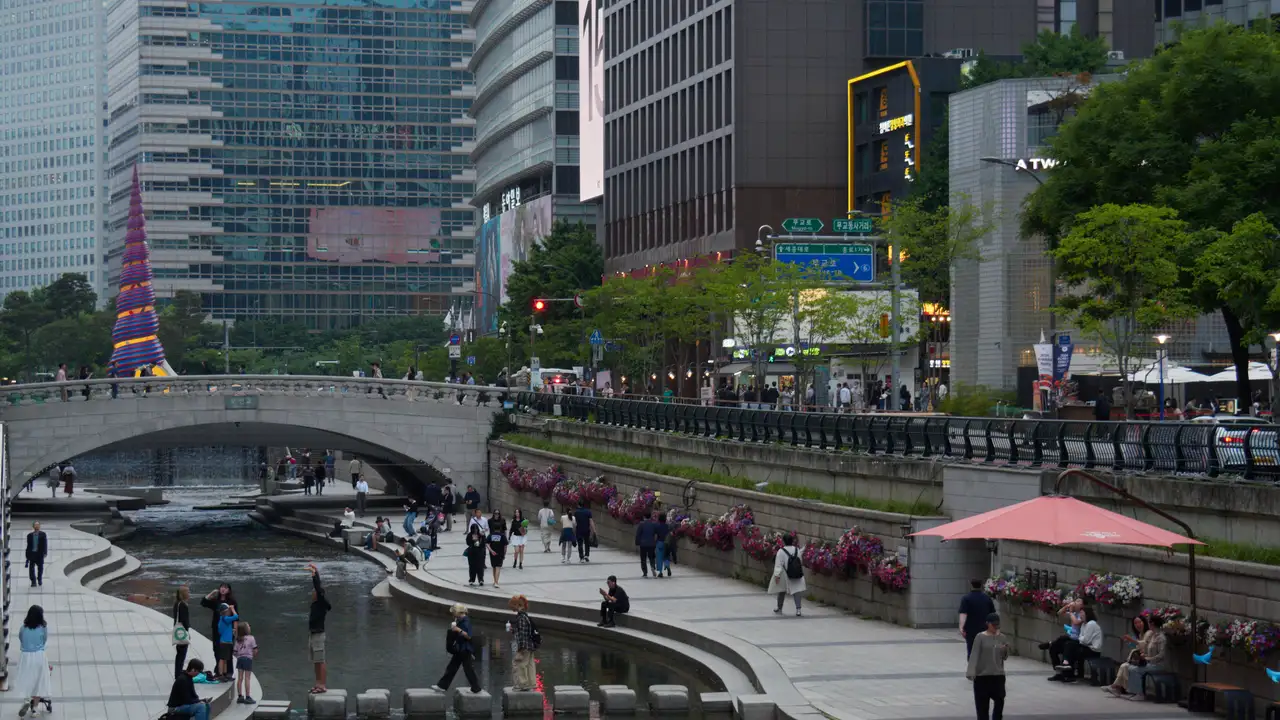 People walking along the Cheonggyecheon stream path in central Seoul at dusk, with a stone bridge and office towers behind