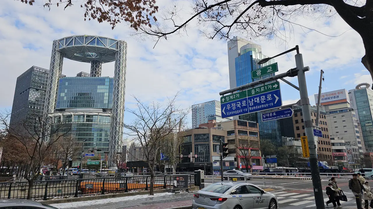 A downtown Seoul intersection with a road sign pointing to Ujeongguk-ro and Namdaemun-ro in Korean and English, high-rise buildings behind