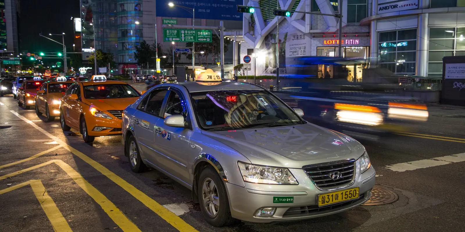 Taxis queued along a kerb in central Seoul at night, a silver sedan with a yellow plate in front and orange cabs behind it with red 빈차 vacancy signs lit in their windscreens