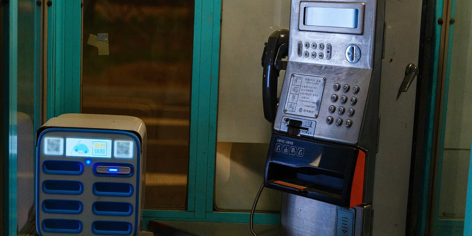 A coin-and-card payphone inside a Korean phone booth, next to a power bank rental kiosk