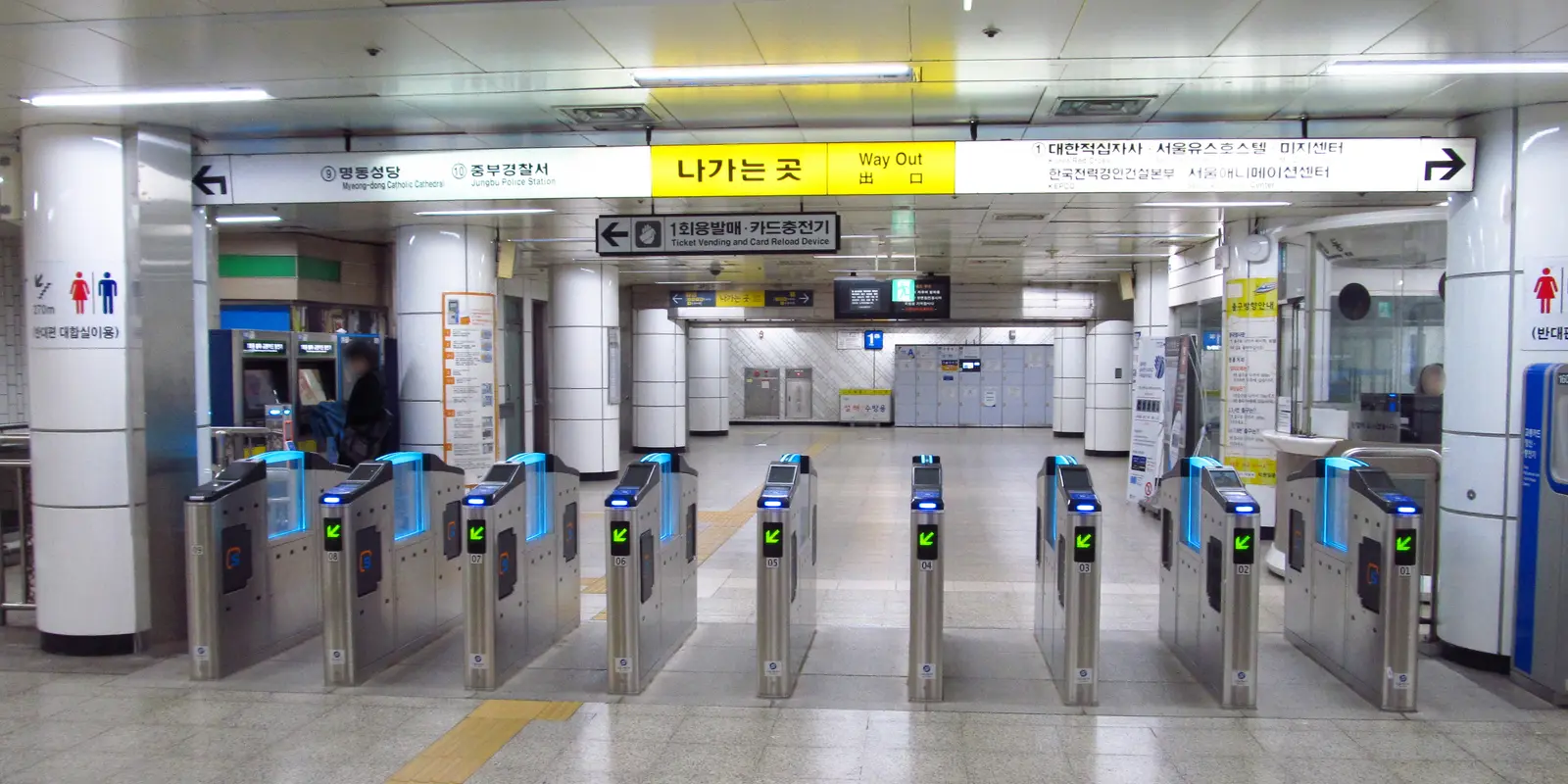 ソウル地下鉄4号線・明洞駅の改札機の列。緑の矢印が点灯し、上には黄色い出口表示と、左の券売・チャージ機を示す案内板